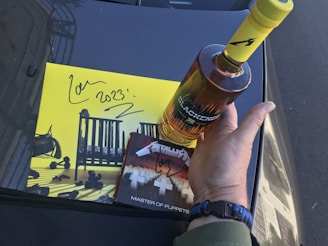 A hand holds a bottle of whiskey labeled 'Blackened', above a car hood. Next to it is a yellow album cover with a drawing of a crib and the text '2023' with a signature. Below the bottle is a CD cover with 'Master of Puppets' text.