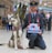 A man in a uniform kneels on the floor of a busy train station next to a large dog. The man is holding a box labeled 'Poppy Appeal' with a red poppy symbol, indicating participation in a charity event. Several people are visible in the background, along with an escalator and directional signs.