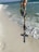A rosary with a crucifix and various decorative beads is held against the backdrop of a sandy beach with gentle ocean waves nearing the shore. The beads are detailed and colorful, displaying a mix of metallic and earthy tones.