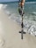 A rosary with a crucifix and various decorative beads is held against the backdrop of a sandy beach with gentle ocean waves nearing the shore. The beads are detailed and colorful, displaying a mix of metallic and earthy tones.