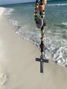 A rosary with a crucifix and various decorative beads is held against the backdrop of a sandy beach with gentle ocean waves nearing the shore. The beads are detailed and colorful, displaying a mix of metallic and earthy tones.