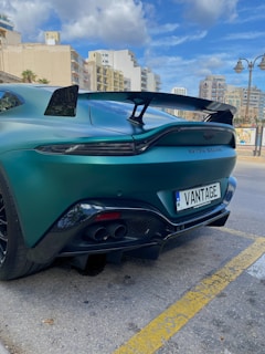 A sleek, green sports car is parked on a city street, showcasing its rear design with a prominent spoiler. The car features dual exhausts and a 'VANTAGE' license plate. Surrounding the car are tall buildings under a partly cloudy sky, with a streetlamp nearby.