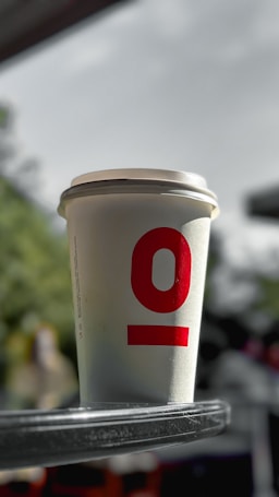 A paper coffee cup with a red logo or number '10' printed on it, placed on a reflective metal surface outdoors. The background is blurred, with hints of greenery and structures.