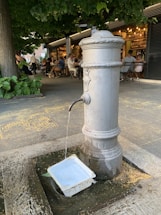 A classic drinking fountain is placed in an outdoor setting with surrounding greenery and a bustling café in the background. Water flows steadily from the spout into a plastic container on the ground. Several people are seated at tables, enjoying their time under what seems to be a tree canopy.
