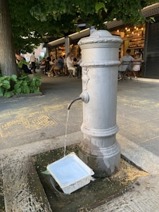A classic drinking fountain is placed in an outdoor setting with surrounding greenery and a bustling café in the background. Water flows steadily from the spout into a plastic container on the ground. Several people are seated at tables, enjoying their time under what seems to be a tree canopy.