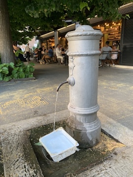 A classic drinking fountain is placed in an outdoor setting with surrounding greenery and a bustling café in the background. Water flows steadily from the spout into a plastic container on the ground. Several people are seated at tables, enjoying their time under what seems to be a tree canopy.