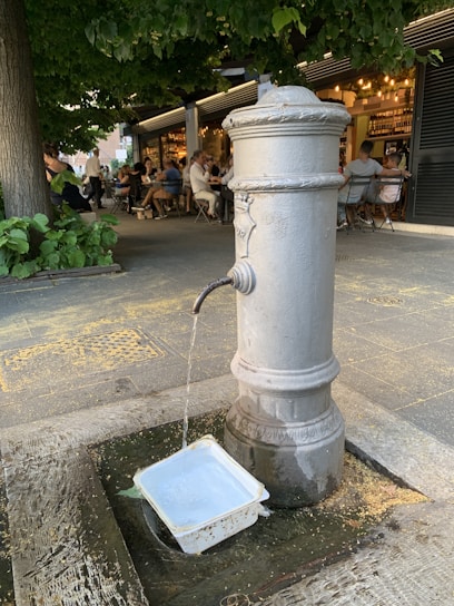 A classic drinking fountain is placed in an outdoor setting with surrounding greenery and a bustling café in the background. Water flows steadily from the spout into a plastic container on the ground. Several people are seated at tables, enjoying their time under what seems to be a tree canopy.