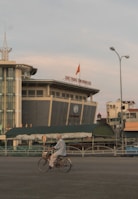 An elderly person is riding a bicycle in an urban area with a large, modern building in the background. The sky is overcast with muted colors.