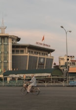 An elderly person is riding a bicycle in an urban area with a large, modern building in the background. The sky is overcast with muted colors.