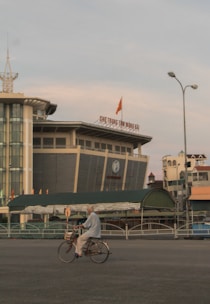 An elderly person is riding a bicycle in an urban area with a large, modern building in the background. The sky is overcast with muted colors.