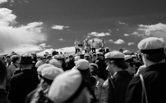 A large gathering of people wearing uniform hats, possibly a celebration or event. In the background, three individuals perform on musical instruments, including a trombone and a drum, standing on an elevated platform under a partly cloudy sky.