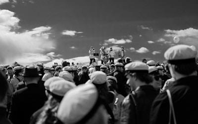 A large gathering of people wearing uniform hats, possibly a celebration or event. In the background, three individuals perform on musical instruments, including a trombone and a drum, standing on an elevated platform under a partly cloudy sky.