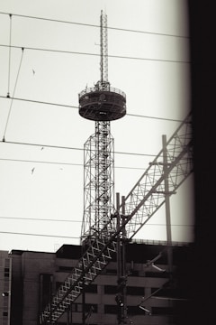 A tall communication tower stands against the sky, featuring a lattice structure and circular platform near the top. Several birds are flying nearby, adding a sense of motion. Power lines and parts of other structures intersect in front of the tower, creating a complex composition of lines and angles.