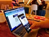 A person holds a smartphone in their hand, scanning a QR code displayed on a laptop screen. The setting appears to be a cafe with people sitting in the background. Nearby, there's an open notebook or diary on the table.