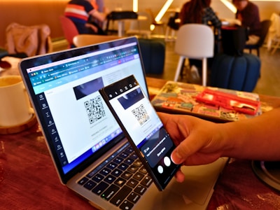 A person holds a smartphone in their hand, scanning a QR code displayed on a laptop screen. The setting appears to be a cafe with people sitting in the background. Nearby, there's an open notebook or diary on the table.