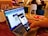 A person holds a smartphone in their hand, scanning a QR code displayed on a laptop screen. The setting appears to be a cafe with people sitting in the background. Nearby, there's an open notebook or diary on the table.