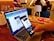 A person holds a smartphone in their hand, scanning a QR code displayed on a laptop screen. The setting appears to be a cafe with people sitting in the background. Nearby, there's an open notebook or diary on the table.