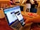 A person holds a smartphone in their hand, scanning a QR code displayed on a laptop screen. The setting appears to be a cafe with people sitting in the background. Nearby, there's an open notebook or diary on the table.