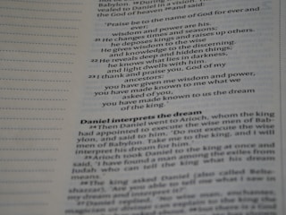 A close-up of a Bible page with text in two columns. It includes verses and a heading 'Daniel interprets the dream'. The text is printed in a serif font, and there are chapter and verse numbers along the margins. The page has a beige tint.