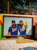 A vibrant mural painted on an outdoor wall depicts two cricket players in blue jerseys. The players are wearing caps and one is holding a cricket bat. The scene is lit by artificial lighting and is surrounded by a border of colorful patterns.