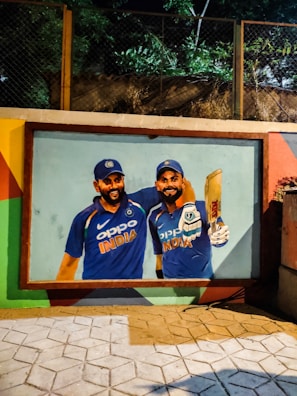 A vibrant mural painted on an outdoor wall depicts two cricket players in blue jerseys. The players are wearing caps and one is holding a cricket bat. The scene is lit by artificial lighting and is surrounded by a border of colorful patterns.