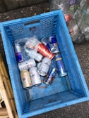 A blue crate filled with various empty aluminum cans, including some from energy drinks and beer. The crate is on a rough-textured ground, surrounded by other containers and bags filled with recyclable materials. The cans are mostly silver with distinctive brand designs.