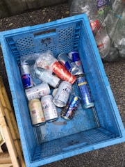A blue crate filled with various empty aluminum cans, including some from energy drinks and beer. The crate is on a rough-textured ground, surrounded by other containers and bags filled with recyclable materials. The cans are mostly silver with distinctive brand designs.