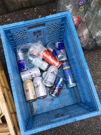A blue crate filled with various empty aluminum cans, including some from energy drinks and beer. The crate is on a rough-textured ground, surrounded by other containers and bags filled with recyclable materials. The cans are mostly silver with distinctive brand designs.
