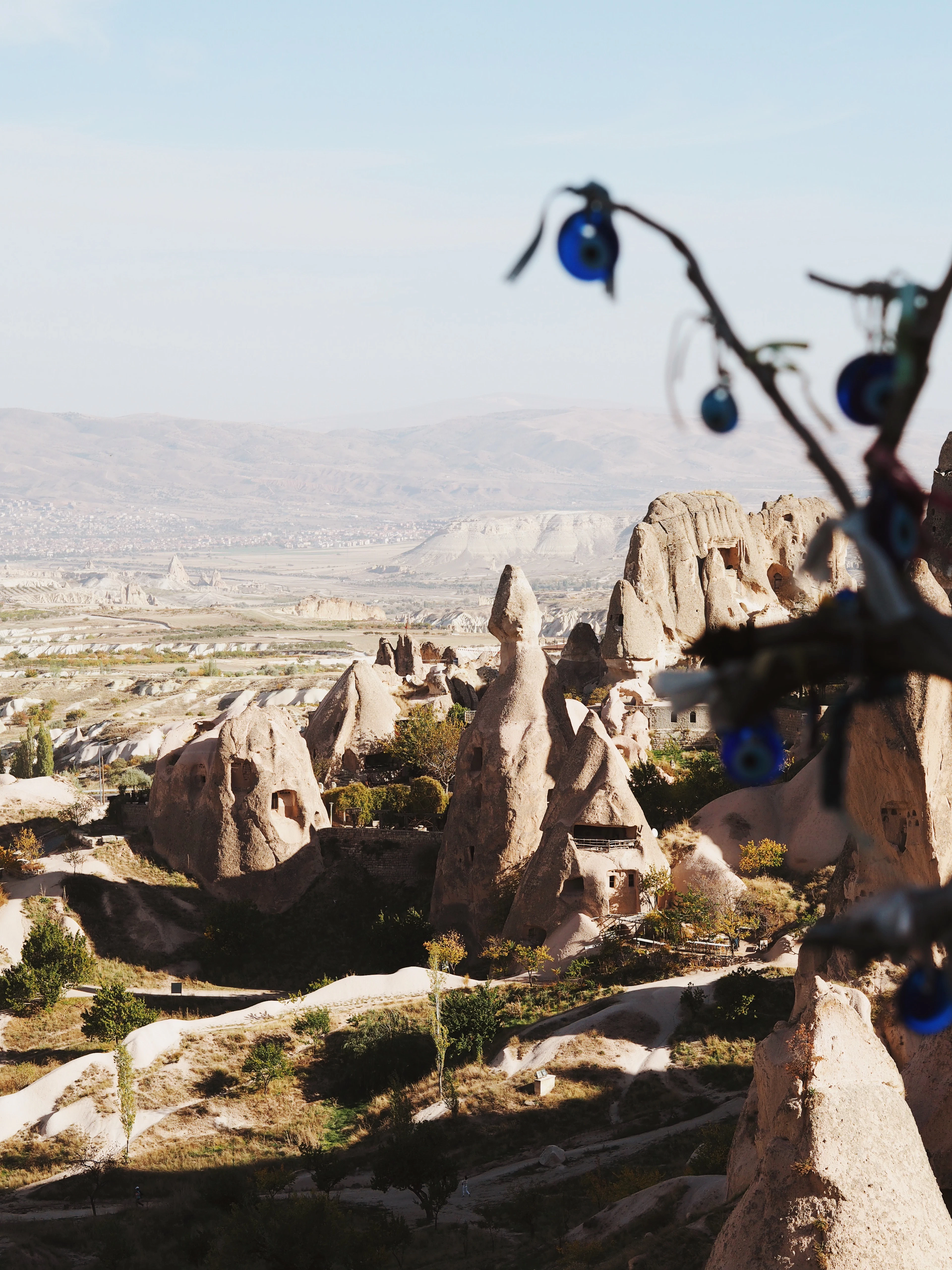 Blue nazar evil-eye amulets hanging from a branch frame a valley of fairy chimneys below.