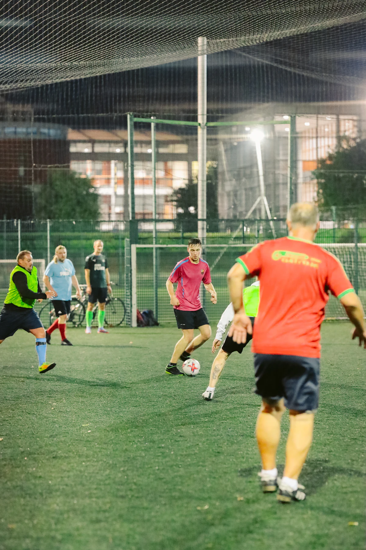 Players of all ages in a floodlit small sided game inside a fenced cage
