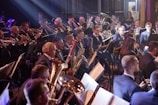 A large group of musicians is playing in an orchestra setting. Many are playing brass instruments such as trombones, trumpets, and tubas, while others are playing woodwind instruments like clarinets and saxophones. The musicians are seated in rows, dressed in formal attire including suits and bow ties, focused on their music. The setting appears to be a stage with dim lighting, and sheet music is visible on stands in front of them.