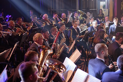 A large group of musicians is playing in an orchestra setting. Many are playing brass instruments such as trombones, trumpets, and tubas, while others are playing woodwind instruments like clarinets and saxophones. The musicians are seated in rows, dressed in formal attire including suits and bow ties, focused on their music. The setting appears to be a stage with dim lighting, and sheet music is visible on stands in front of them.
