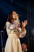 A woman is singing into a microphone on stage, wearing a long beige dress with colorful floral embroidery on the sleeves and chest. Her hair is long and brown, and there is a blurred musician in the background.