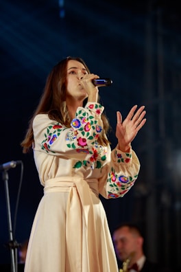 A woman is singing into a microphone on stage, wearing a long beige dress with colorful floral embroidery on the sleeves and chest. Her hair is long and brown, and there is a blurred musician in the background.