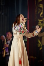 A woman is passionately singing into a microphone while wearing a long, cream-colored dress with colorful floral embroidery on the sleeves. Her posture is expressive and full of emotion, with one hand gripping the microphone and the other raised. In the background, a musician is visible playing a saxophone.