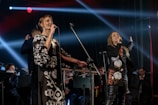 Two performers on stage hold microphones and are passionately singing. They are dressed in stylish black and white outfits with intricate patterns. Behind them, musicians play in a band, softly lit by colorful stage lights and beams.