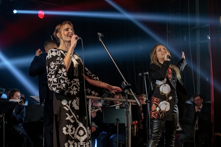 Two performers on stage hold microphones and are passionately singing. They are dressed in stylish black and white outfits with intricate patterns. Behind them, musicians play in a band, softly lit by colorful stage lights and beams.