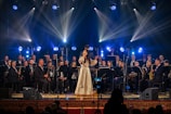 A woman in a long, traditional dress sings on stage, surrounded by a large orchestra of musicians playing various instruments. Bright spotlights and colored stage lights illuminate the performers, creating a vibrant atmosphere.