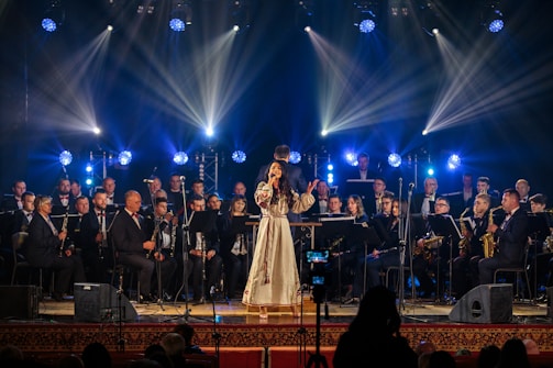A woman in a long, traditional dress sings on stage, surrounded by a large orchestra of musicians playing various instruments. Bright spotlights and colored stage lights illuminate the performers, creating a vibrant atmosphere.