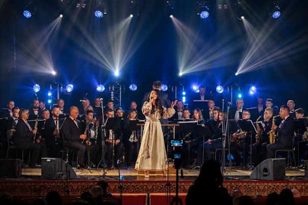 A woman in a long, traditional dress sings on stage, surrounded by a large orchestra of musicians playing various instruments. Bright spotlights and colored stage lights illuminate the performers, creating a vibrant atmosphere.