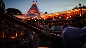 A carnival-style amusement ride featuring colorful cars shaped like elephants illuminated by warm carnival lights. A tent-like structure with signage glows in the evening light, and people gather around enjoying the festive atmosphere.