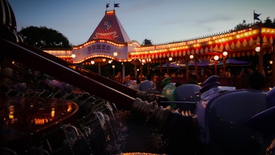 A carnival-style amusement ride featuring colorful cars shaped like elephants illuminated by warm carnival lights. A tent-like structure with signage glows in the evening light, and people gather around enjoying the festive atmosphere.