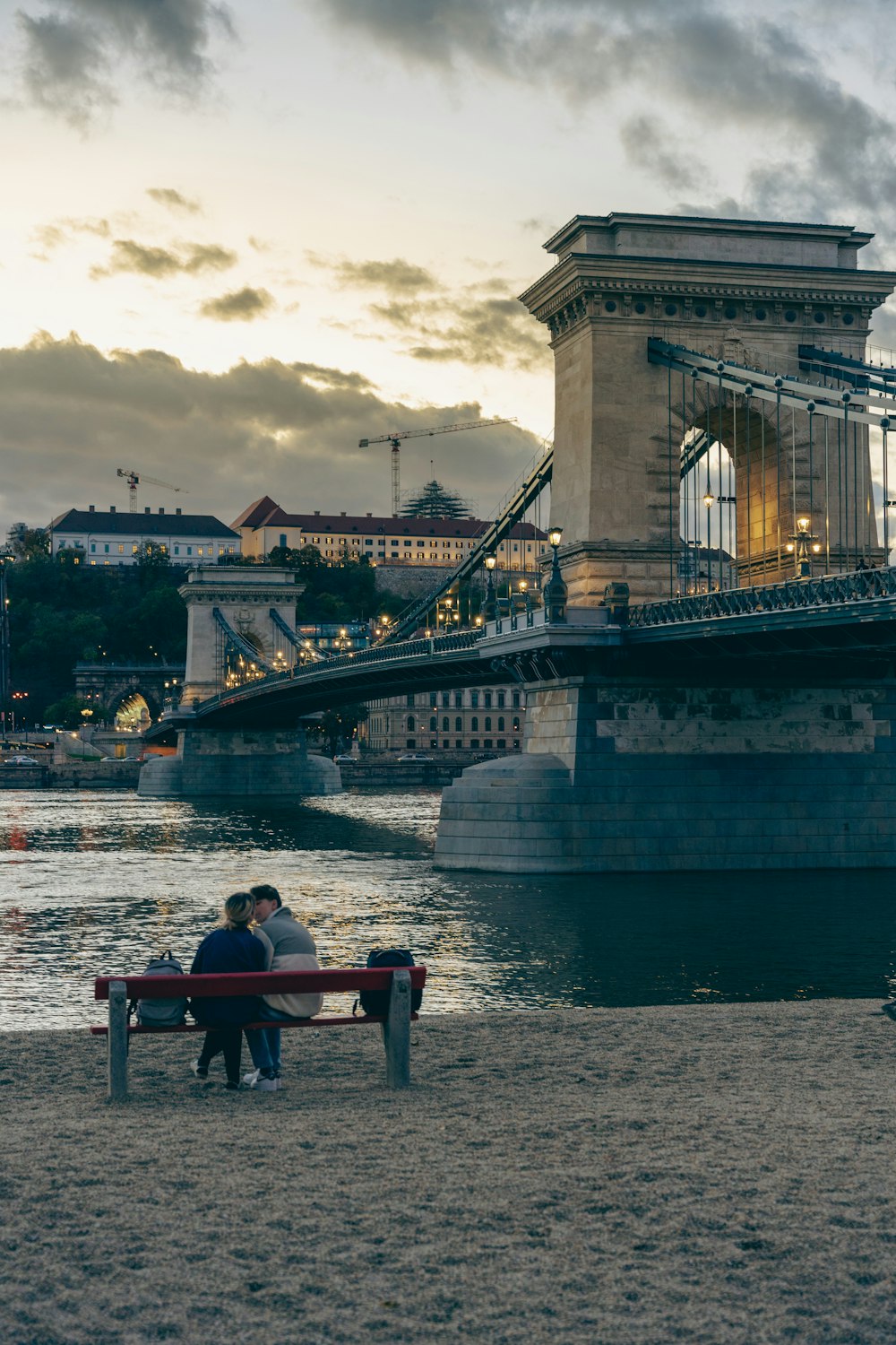 two people sitting on a bench in front of a bridge