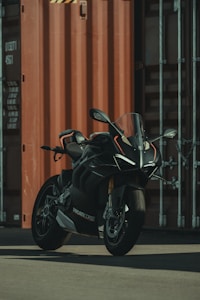 A sleek, modern motorcycle with the brand 'Ducati Corse' visible on its body stands in front of a large shipping container. The motorcycle has a dark, matte finish with subtle orange accents. It is positioned on a flat, paved surface and is well-lit by soft, ambient light.