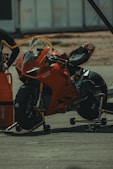 A red sports motorcycle is positioned on a stand in a pit stop area. There's a fuel hose connected to the motorcycle, indicating it is being refueled or serviced. The background includes a blurred view of other pit stop equipment and structures.