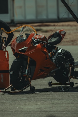 A red sports motorcycle is positioned on a stand in a pit stop area. There's a fuel hose connected to the motorcycle, indicating it is being refueled or serviced. The background includes a blurred view of other pit stop equipment and structures.