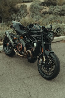 A sleek, modern motorcycle with a black matte finish and orange accents is parked on a concrete surface. The bike features a detailed exhaust system and large tires, with a style suitable for high performance. The background consists of a blurred, greenery-covered hillside.