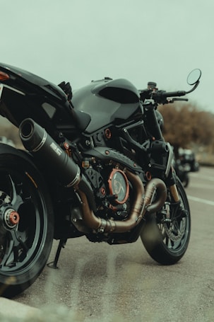 A sleek motorcycle is parked on the side of a road, showcasing its exhaust system, engine details, and stylish design. The vehicle has a matte black finish with orange accents and appears to be in excellent condition. The background is slightly blurred, indicating an outdoor setting.