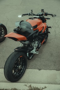 A sleek, modern motorcycle with an aerodynamic design and a bold orange finish. It is parked on a concrete surface, with a black helmet placed on the rear seat. The motorcycle features advanced handlebars and control systems, highlighting its sporty features. Its large rear tire and dual exhaust add to its powerful appearance.