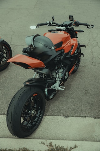 A sleek, modern motorcycle with an aerodynamic design and a bold orange finish. It is parked on a concrete surface, with a black helmet placed on the rear seat. The motorcycle features advanced handlebars and control systems, highlighting its sporty features. Its large rear tire and dual exhaust add to its powerful appearance.